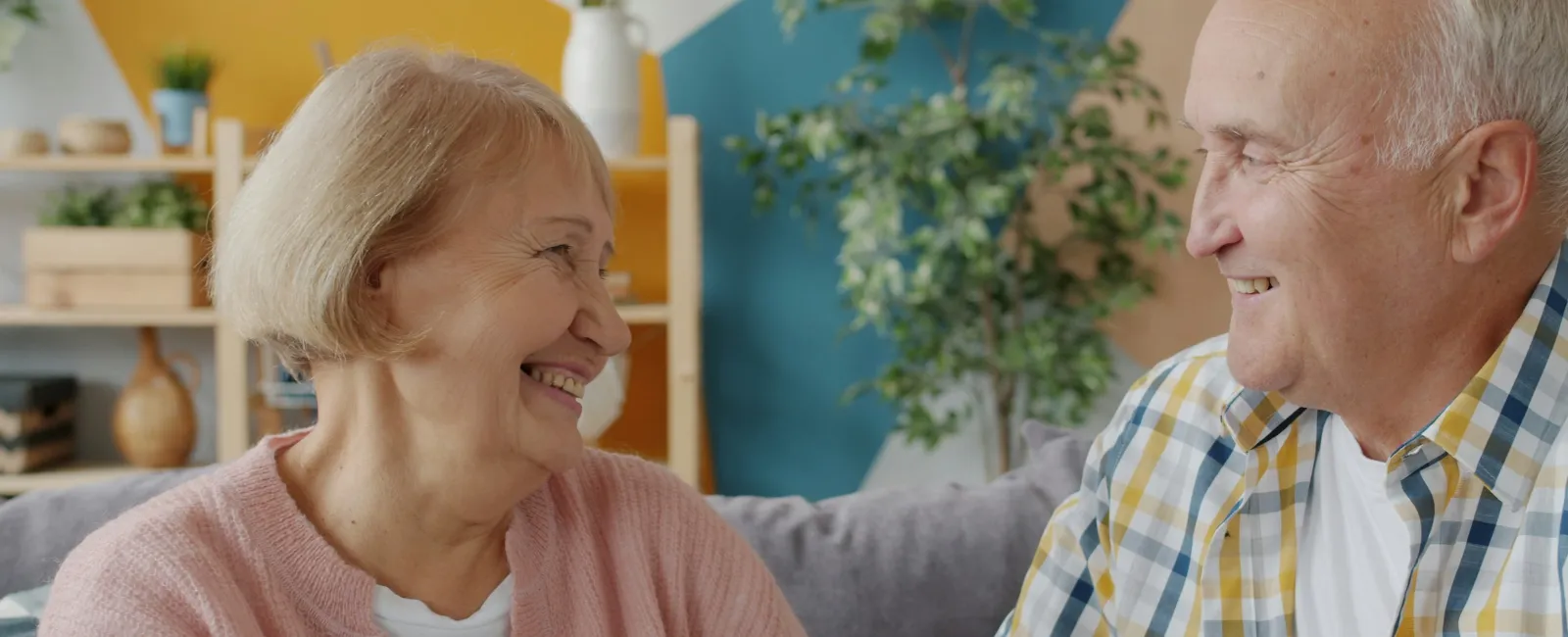 Smiling elderly couple sitting on a couch in a colorful living room, sharing a happy moment together.