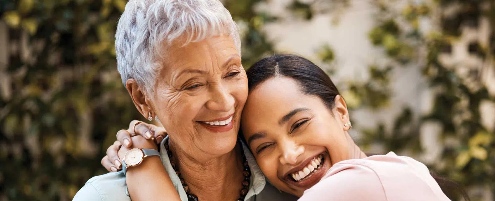 Happy young woman hugging older woman outdoors, both smiling warmly with greenery background.