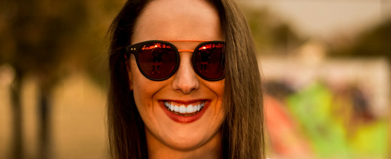 Smiling woman with sunglasses, wearing a light blue tank top, outdoors with a blurred colorful background.