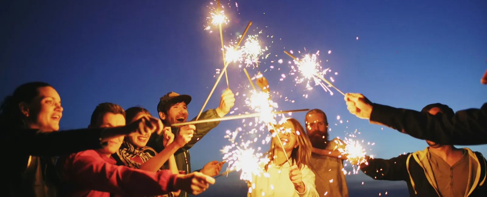 Group of friends joyfully holding sparklers together at dusk near water with city lights in background