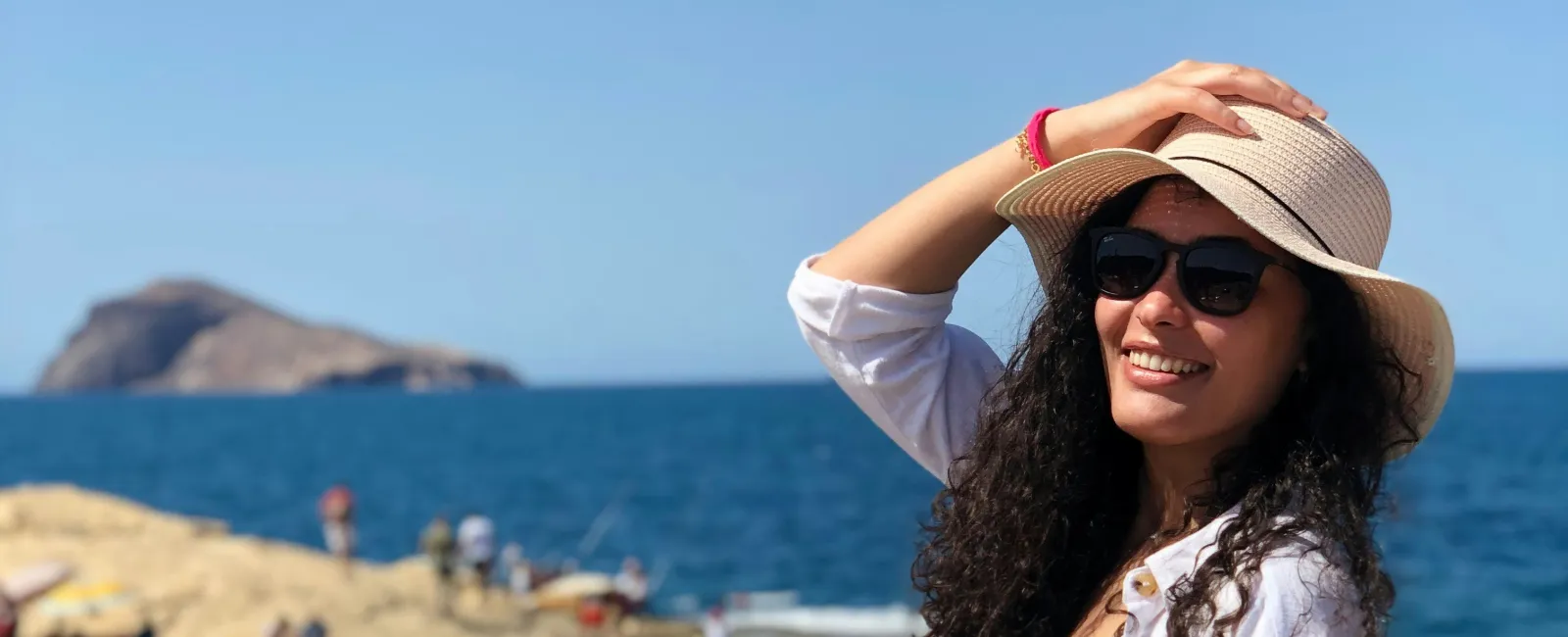 Smiling woman in sunglasses and sunhat stands by rocky seaside with blue ocean and island in background.