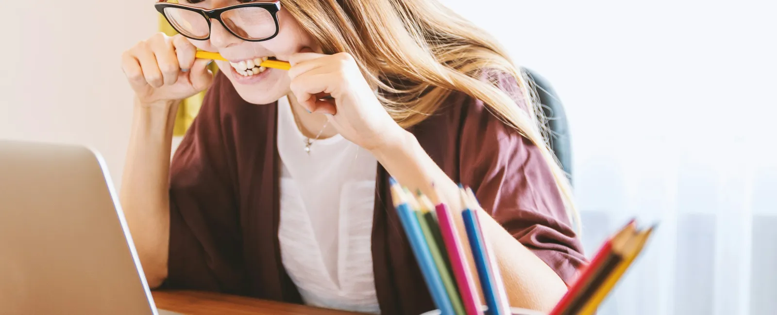 Stressed young woman biting pencil while working on laptop at wooden desk with colored pencils nearby