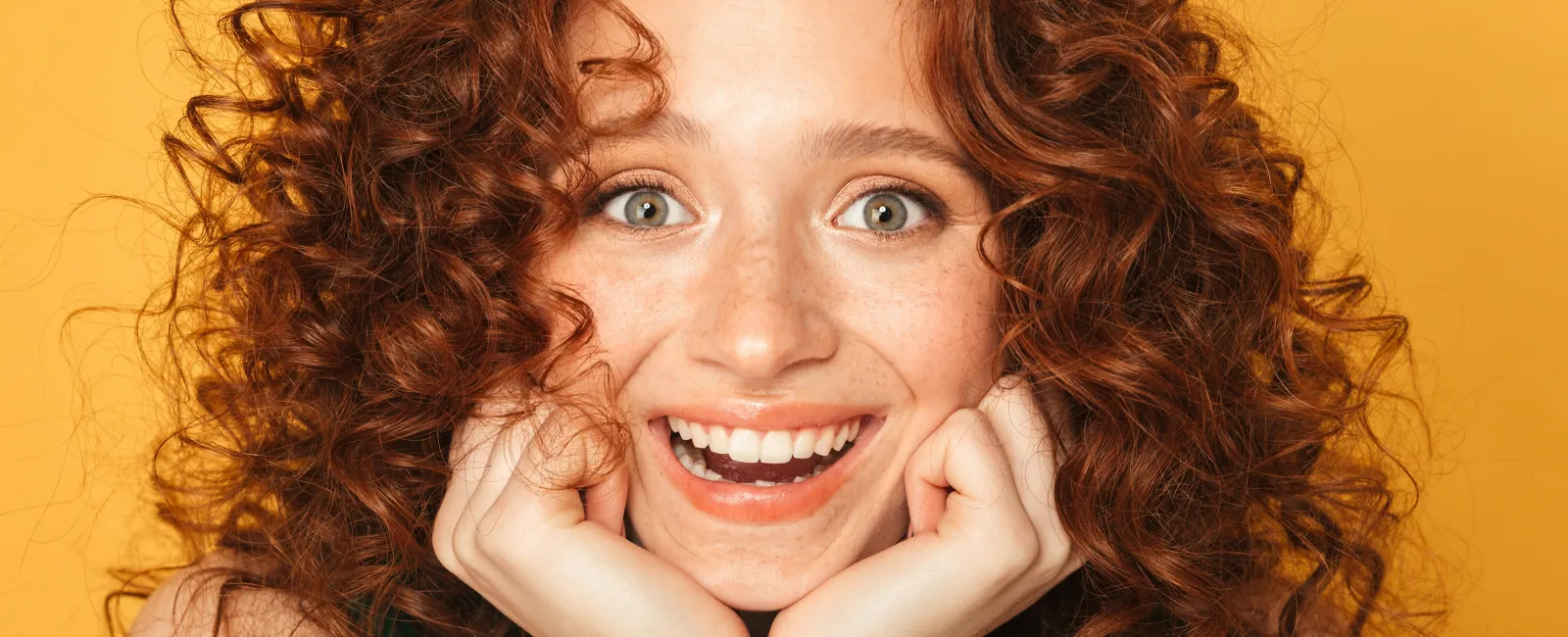 Happy young woman with curly red hair smiling widely against a bright yellow background.