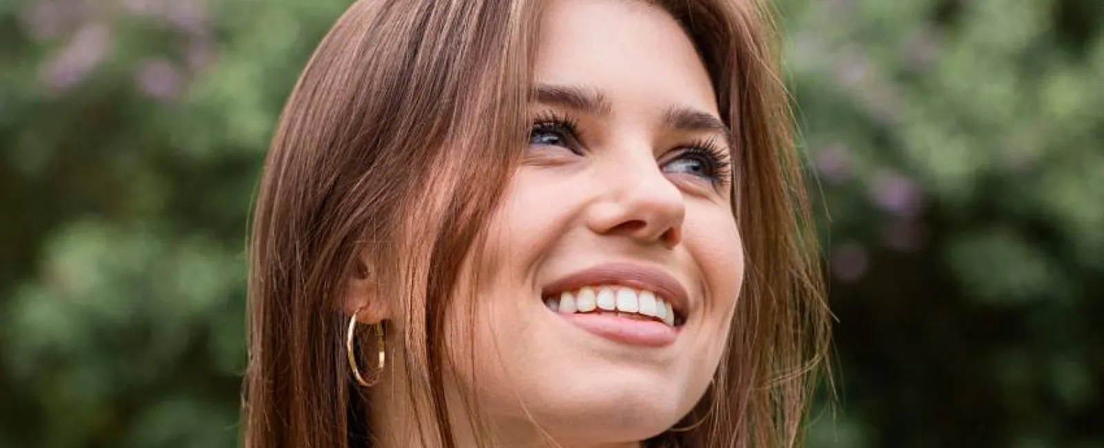 Smiling young woman with brown hair and hoop earrings outdoors with greenery and purple flowers behind her