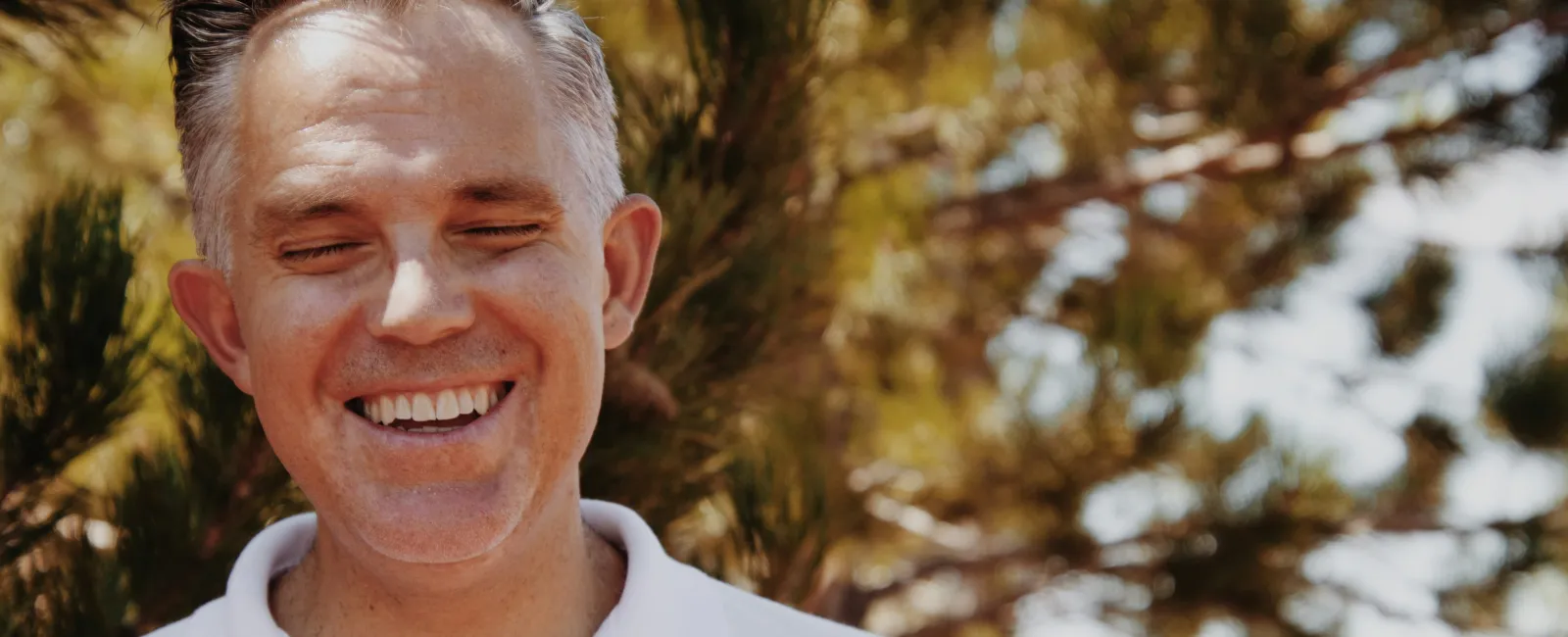 Smiling middle-aged man outdoors with closed eyes and trees in soft focus background under sunlight.