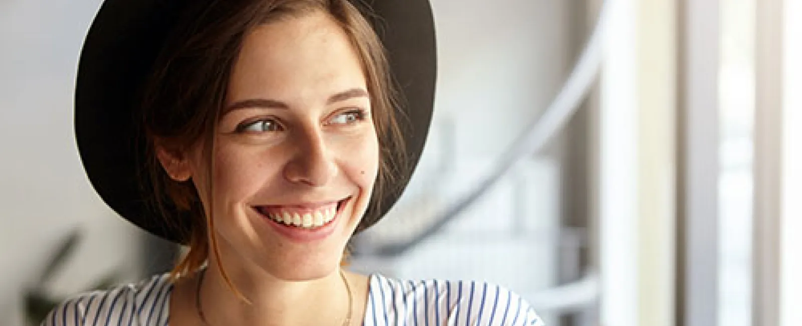 Young woman wearing a hat smiling and holding a smartphone while looking away in a bright indoor setting