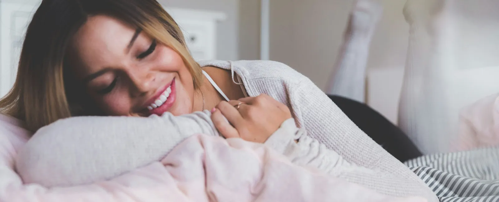 Smiling young woman lying on bed resting on her arm with cozy pillows and casual sweater indoors