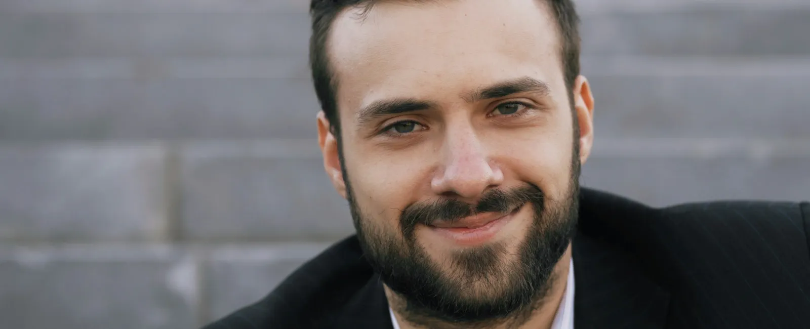 Smiling bearded man in a black suit sitting outdoors with blurred stairs in the background