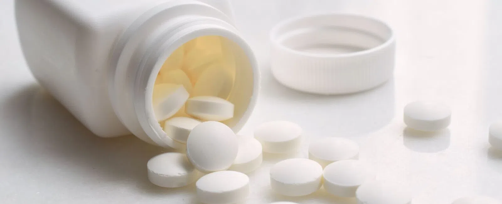 White tablets spilling from an open white plastic pill bottle on a light surface with the bottle cap beside it.