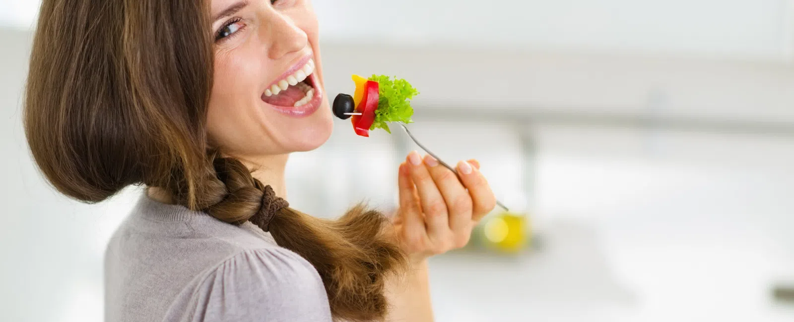 Woman with braided hair happily eating a fresh vegetable salad with olives and lettuce in a bright kitchen.