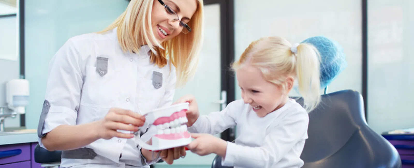 Female dentist teaching little girl about dental care using a teeth model in a bright clinic.
