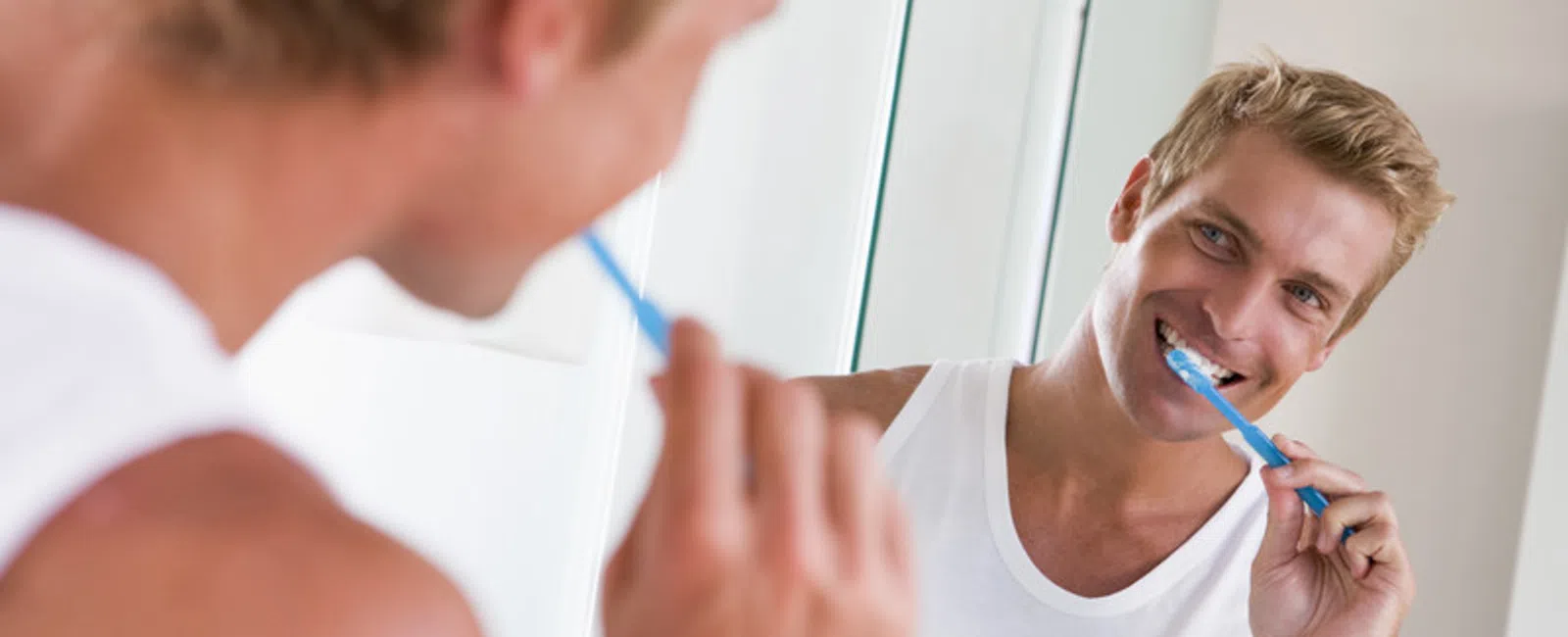 Young man in white tank top brushing his teeth in front of a bathroom mirror smiling.