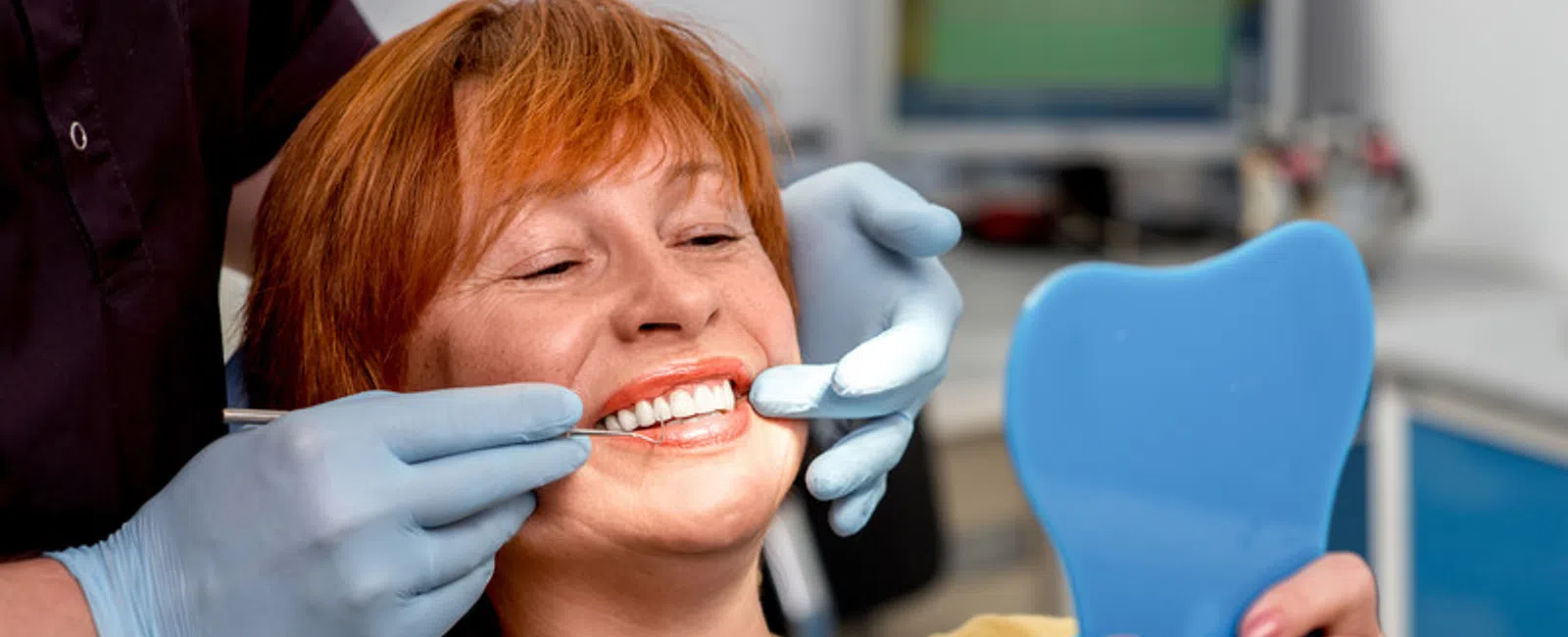 Woman smiling with healthy white teeth during dental checkup, holding a blue tooth-shaped mirror.