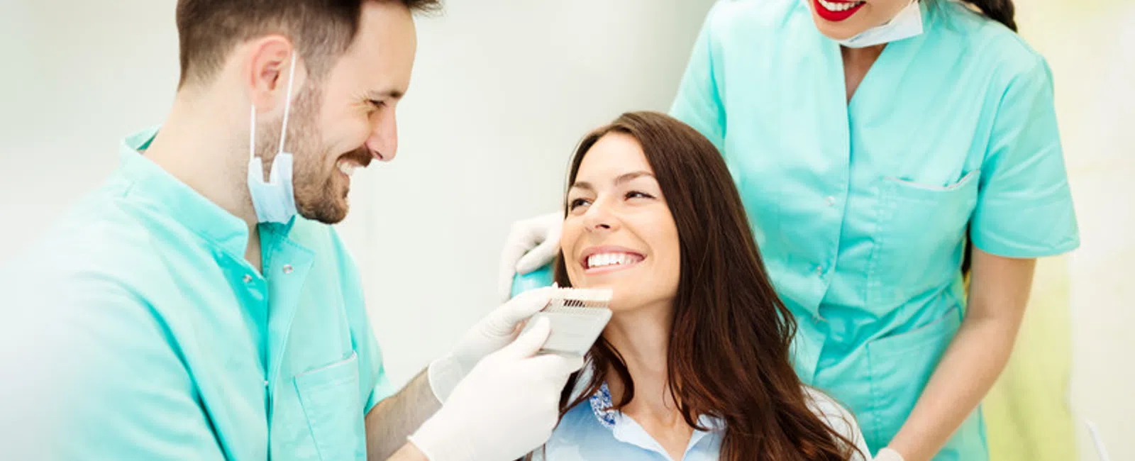 Dentist and assistant smiling while examining a female patient's teeth with a shade guide in a dental clinic.