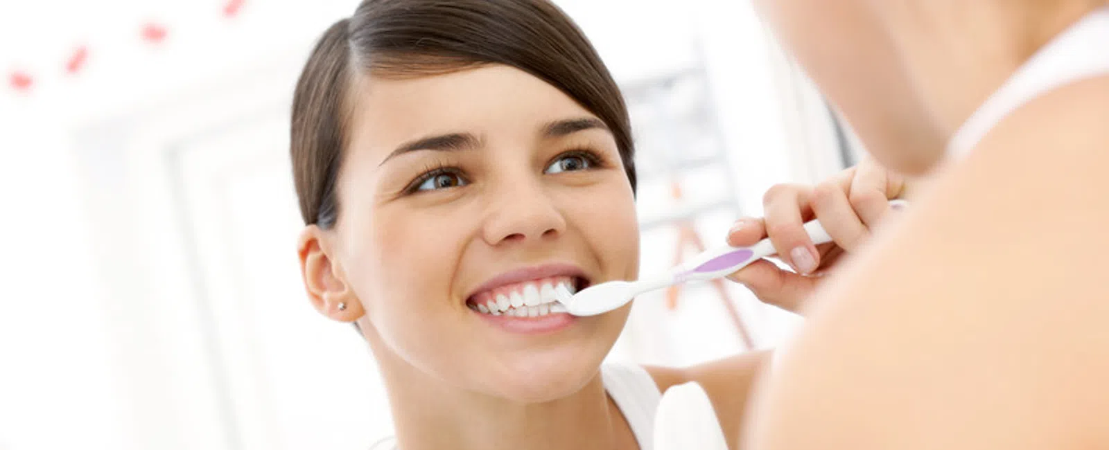 Young woman smiling while brushing her teeth in front of a bathroom mirror with dental products nearby