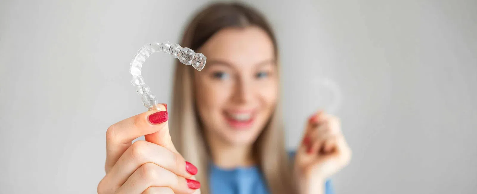 Smiling woman holding clear dental aligners for teeth straightening in a bright, neutral setting.