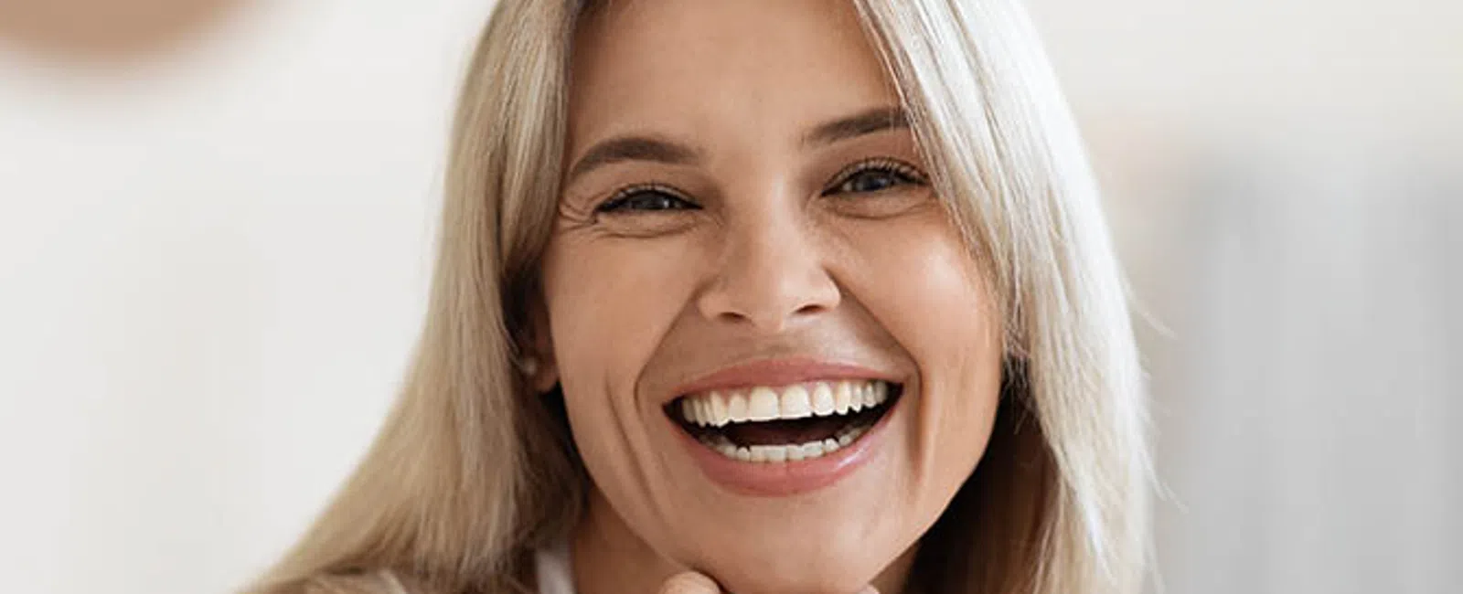 Smiling blonde woman with hand under chin wearing a white blouse in a bright indoor setting