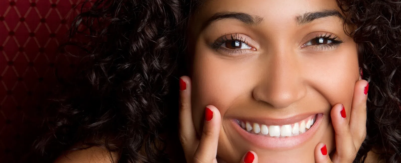 Close-up of a smiling woman with curly hair and red nail polish resting her face in her hands