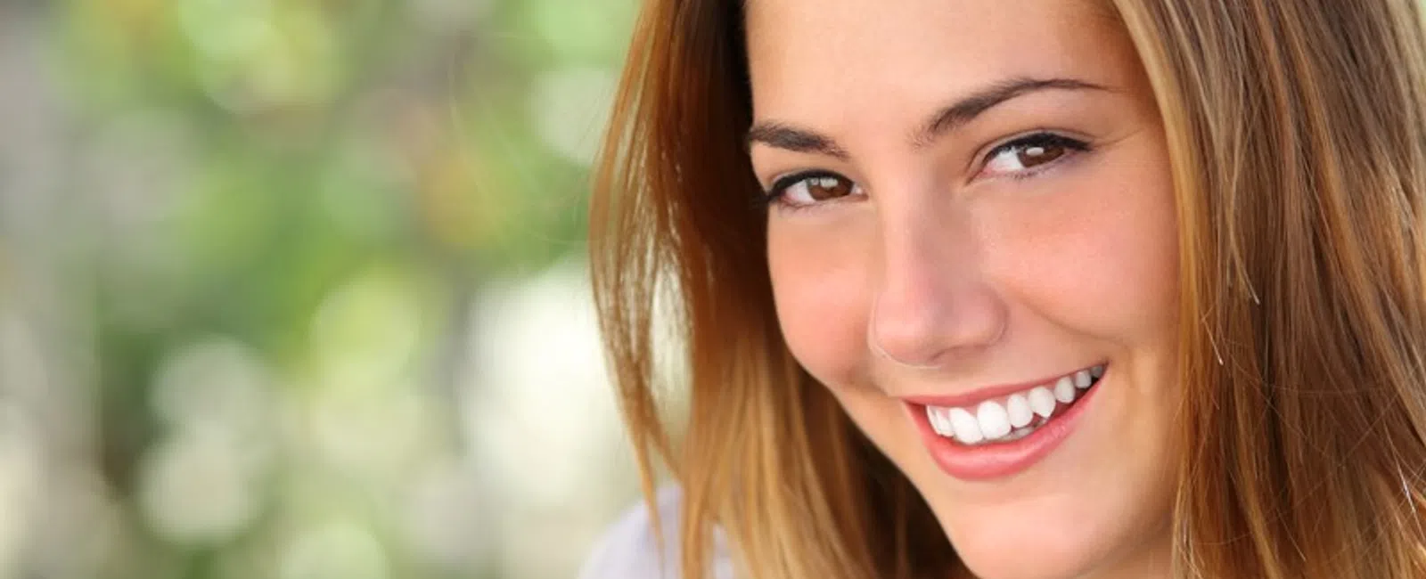 Close-up of a smiling young woman with long light brown hair against a blurred green background