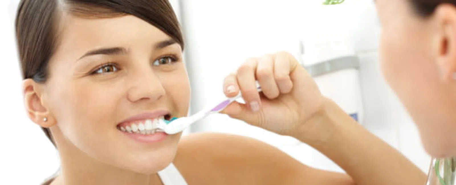 Smiling woman brushing her teeth in front of a bathroom mirror promoting dental hygiene.