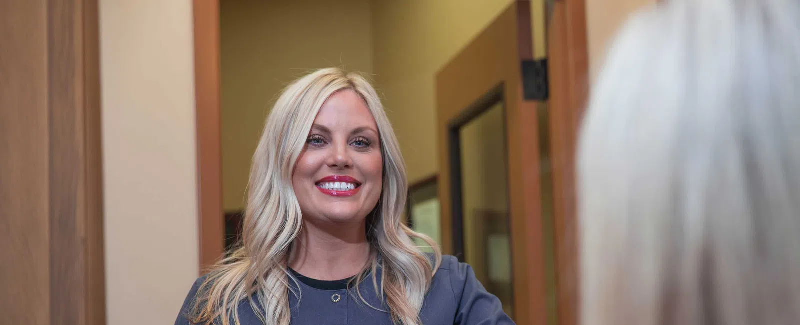 Smiling blonde woman with long hair wearing navy blue scrubs stands behind reception desk in office.