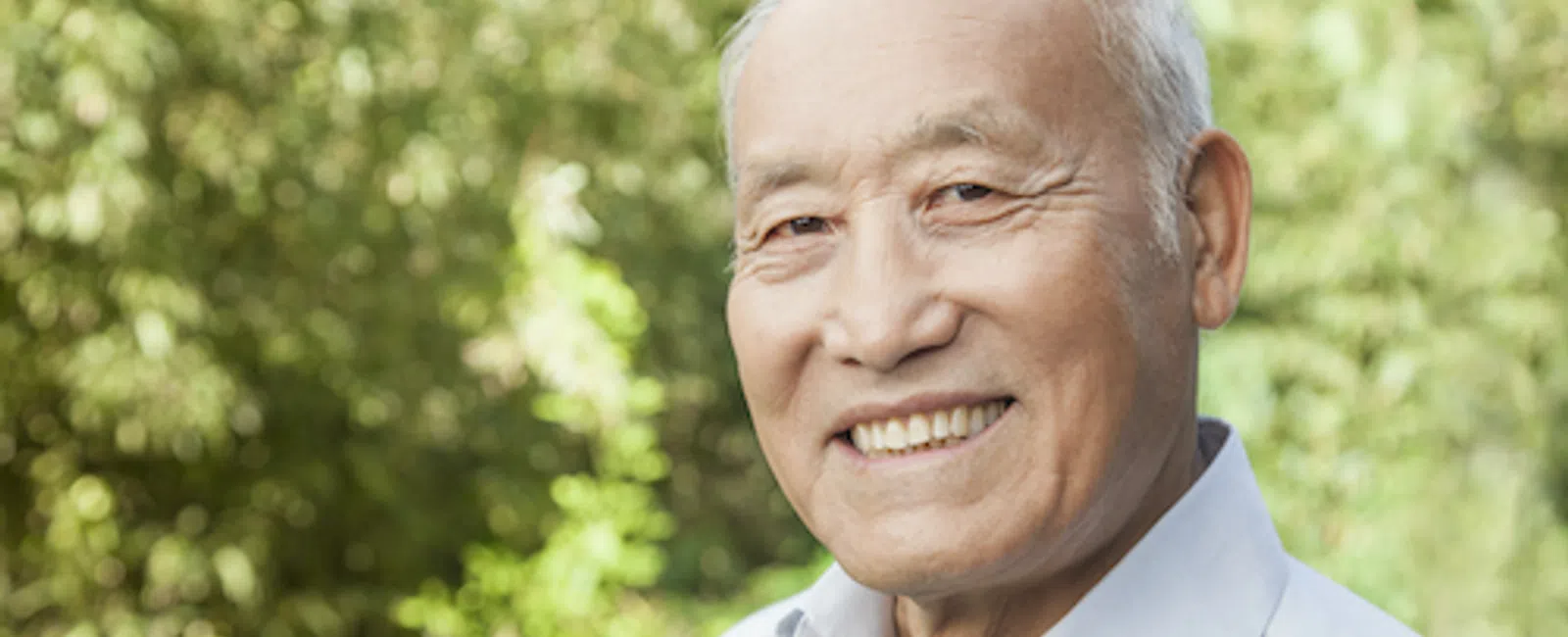 Smiling elderly man with white hair in a light blue shirt standing outdoors with green leafy background