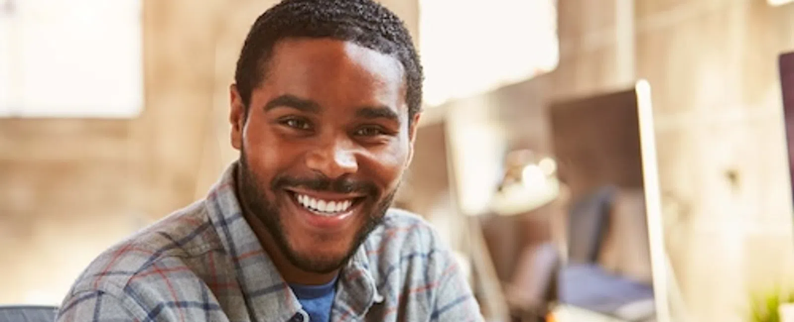 Smiling man wearing plaid shirt sitting at desk in bright modern office workspace with computer in background