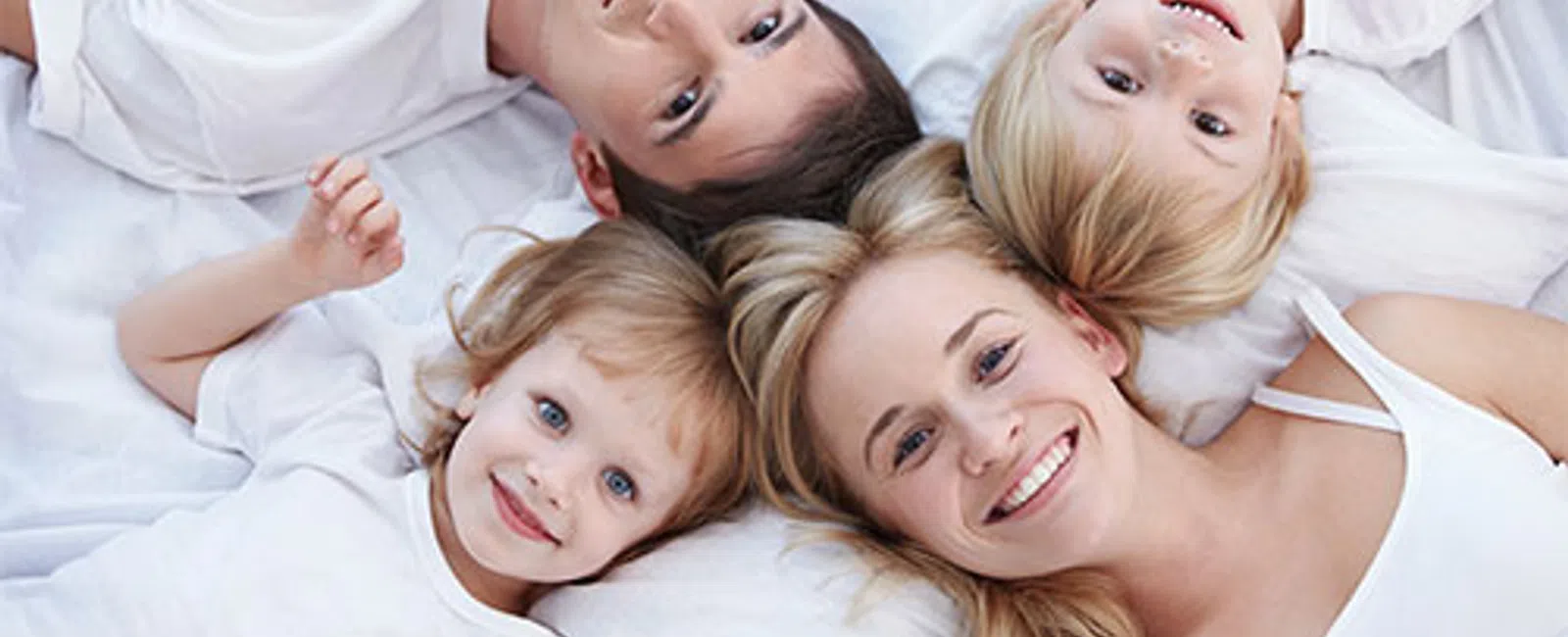 Smiling family of four lying on white bed wearing white clothes looking up at camera.