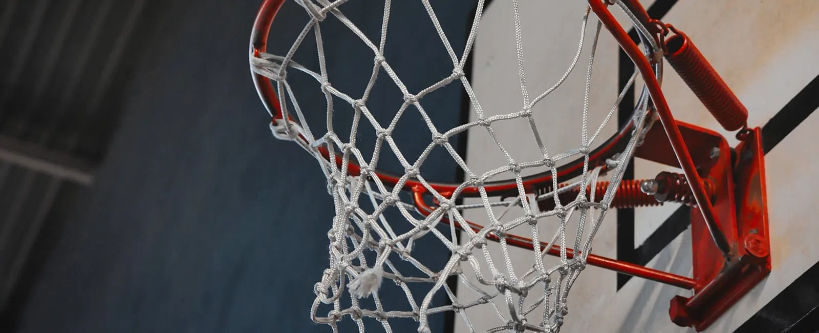 Close-up of a red basketball hoop with white net against a gray and blue indoor gym background.