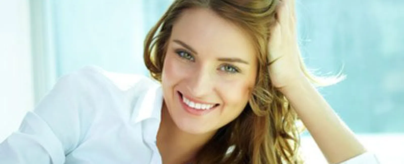 Smiling woman with long hair wearing a white shirt reclining comfortably by a window with natural light.