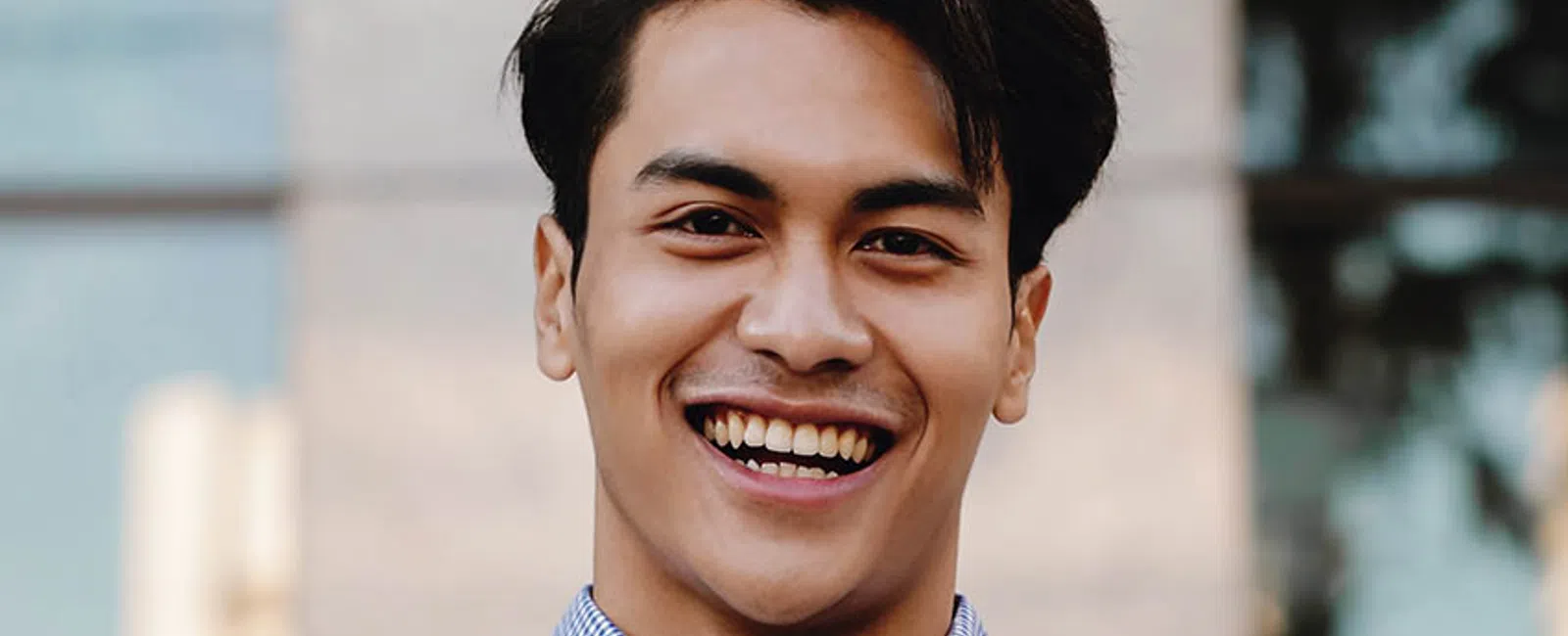 Young man smiling confidently with arms crossed wearing a blue checkered shirt outdoors in urban setting