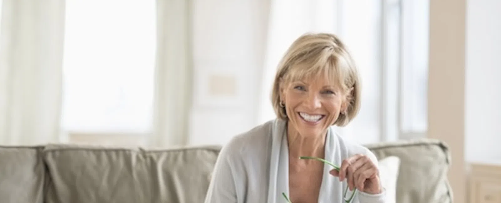 Smiling elderly woman sitting on couch using laptop and holding glasses in bright living room with coffee cup nearby