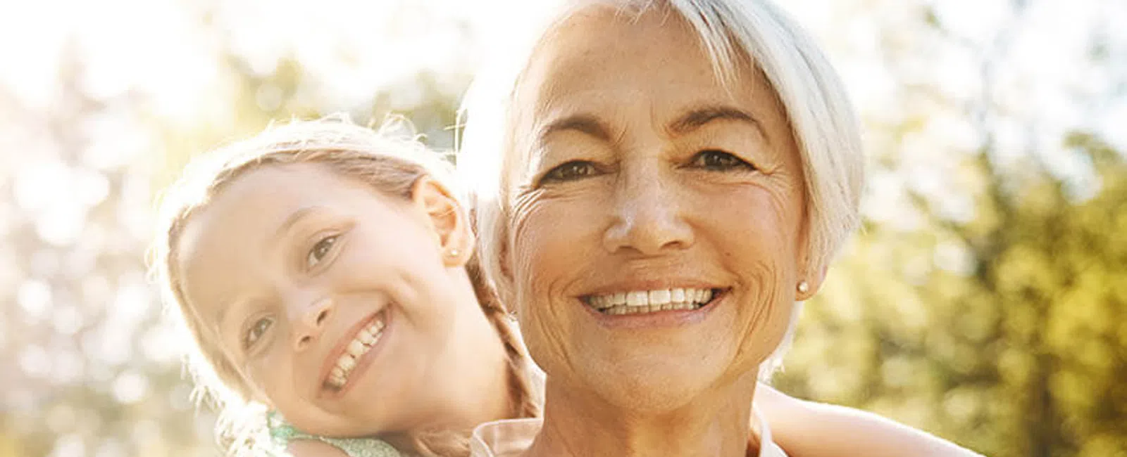 Smiling elderly woman with short white hair carrying a happy young girl on her back outdoors in a sunlit park