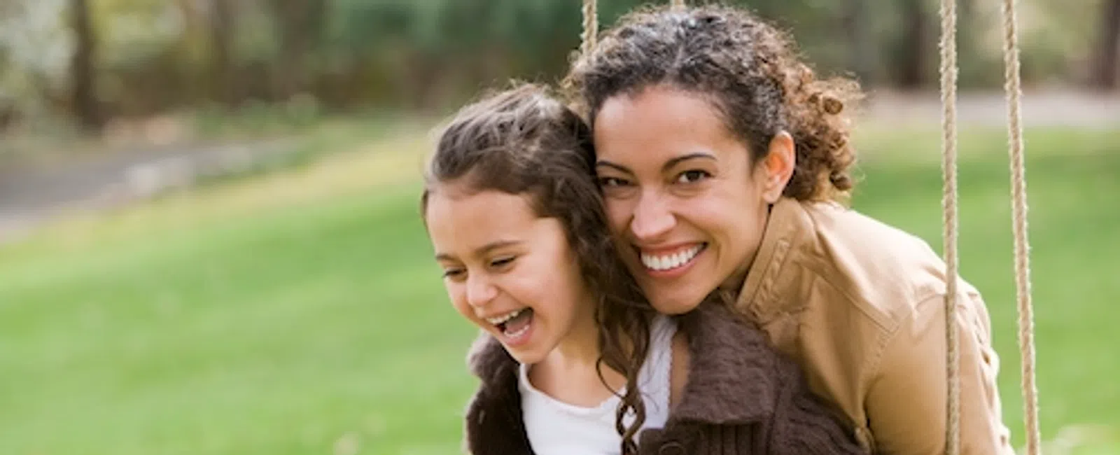 Mother and daughter smiling and enjoying time together on a swing in a green outdoor park setting.