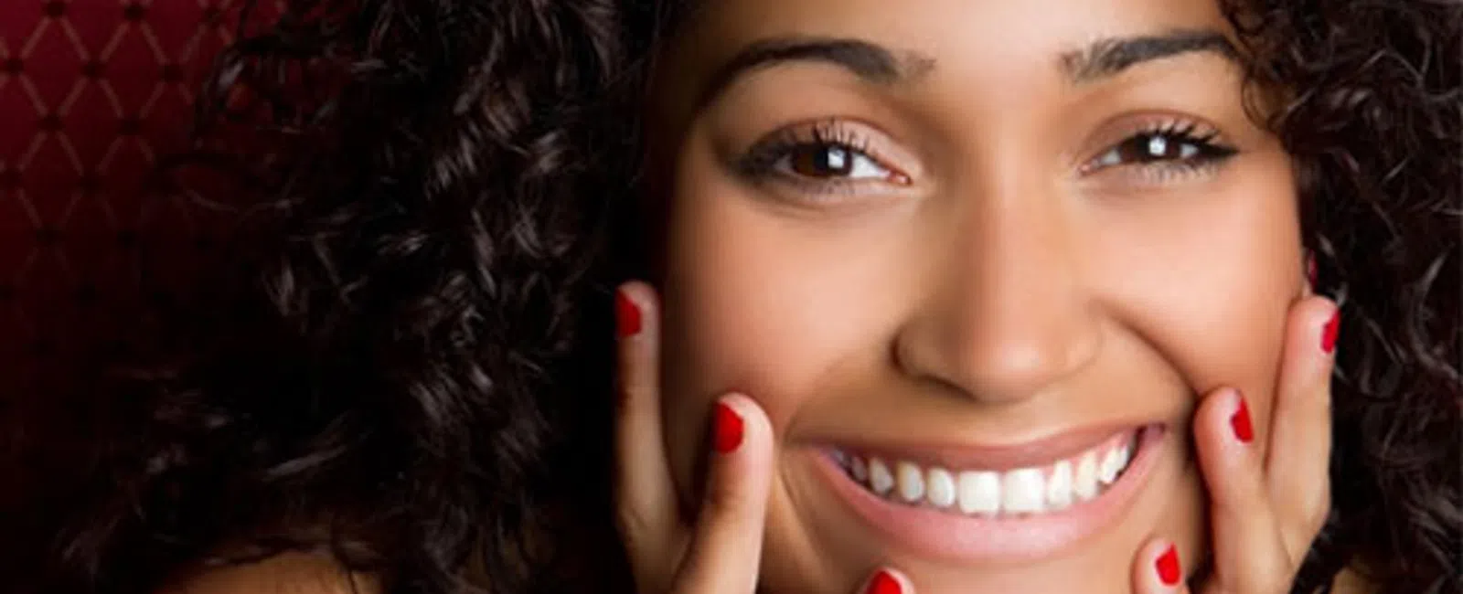 Close-up of a smiling woman with curly hair and red nail polish resting her face in her hands.