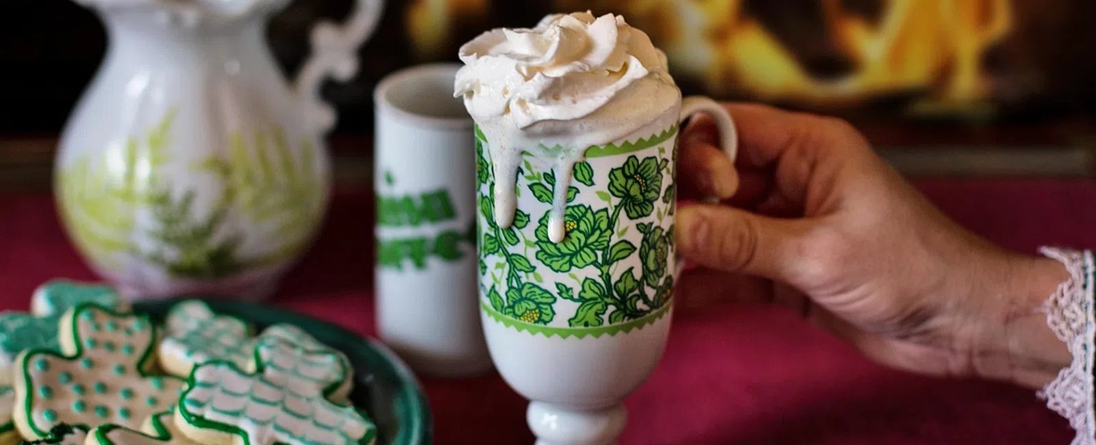 Hand holding vintage green floral mug with whipped cream hot drink, holiday cookies and fireplace in background.