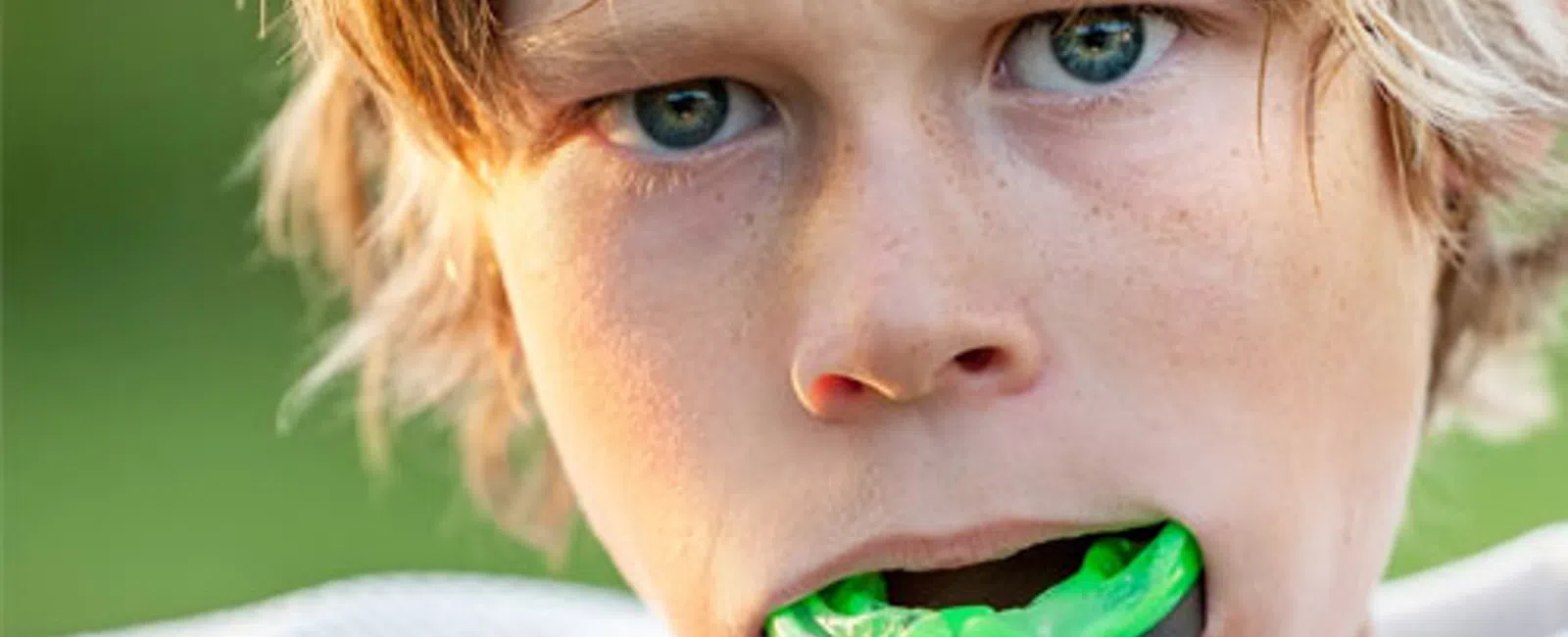 Close-up of a young athlete biting a green mouthguard with focused eyes and sports gear outdoors.