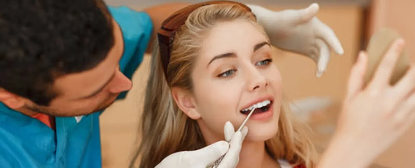Dentist examining a smiling young woman’s teeth while she looks into a handheld mirror during a dental checkup.
