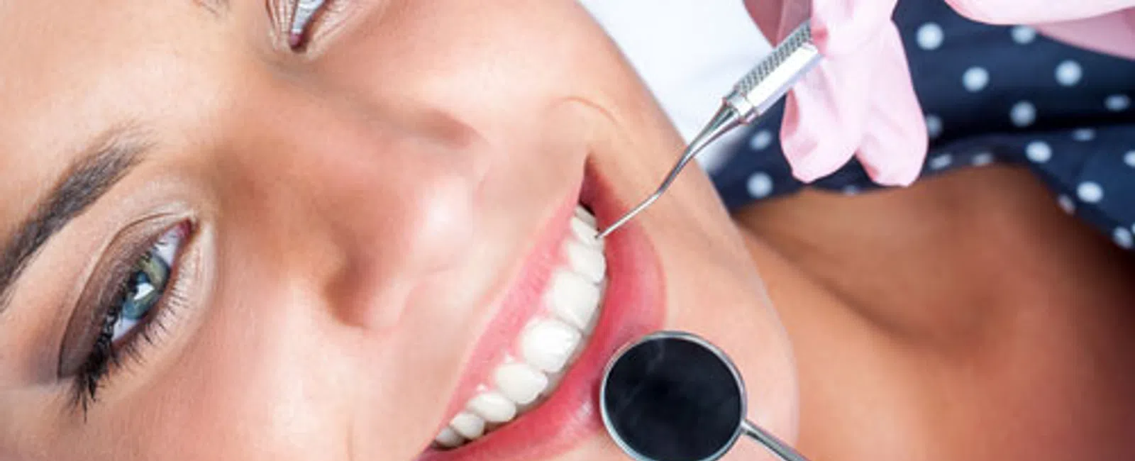 Close-up of a smiling woman undergoing a dental checkup with tools and pink-gloved hands.