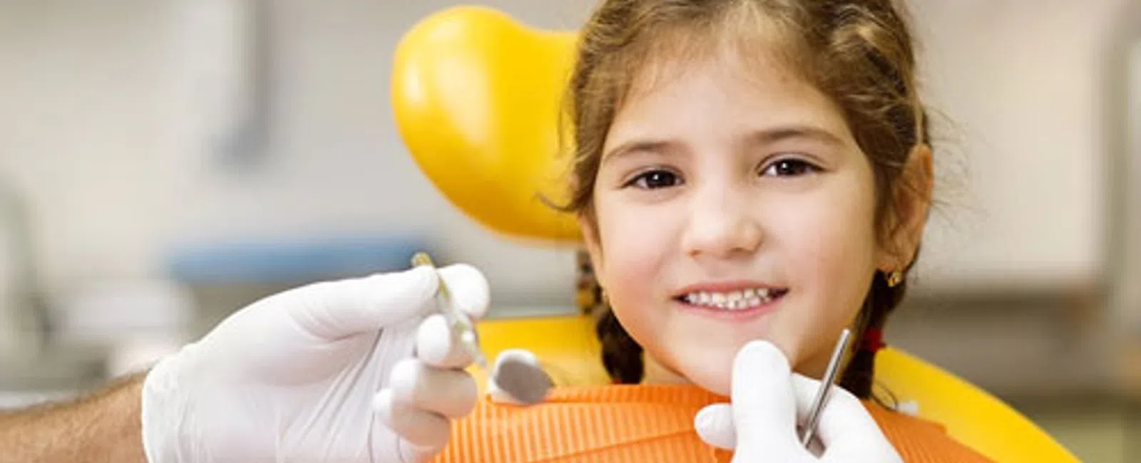 Smiling young girl sitting in a dental chair with dentist's hands holding dental tools nearby.