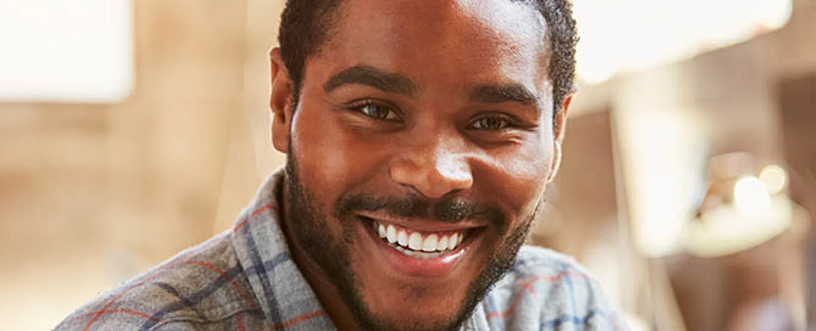Smiling young man wearing a plaid shirt sitting indoors with warm natural light in the background