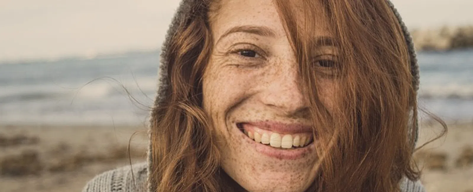 Smiling young woman with red hair and freckles wearing a knitted sweater at the beach on a cloudy day
