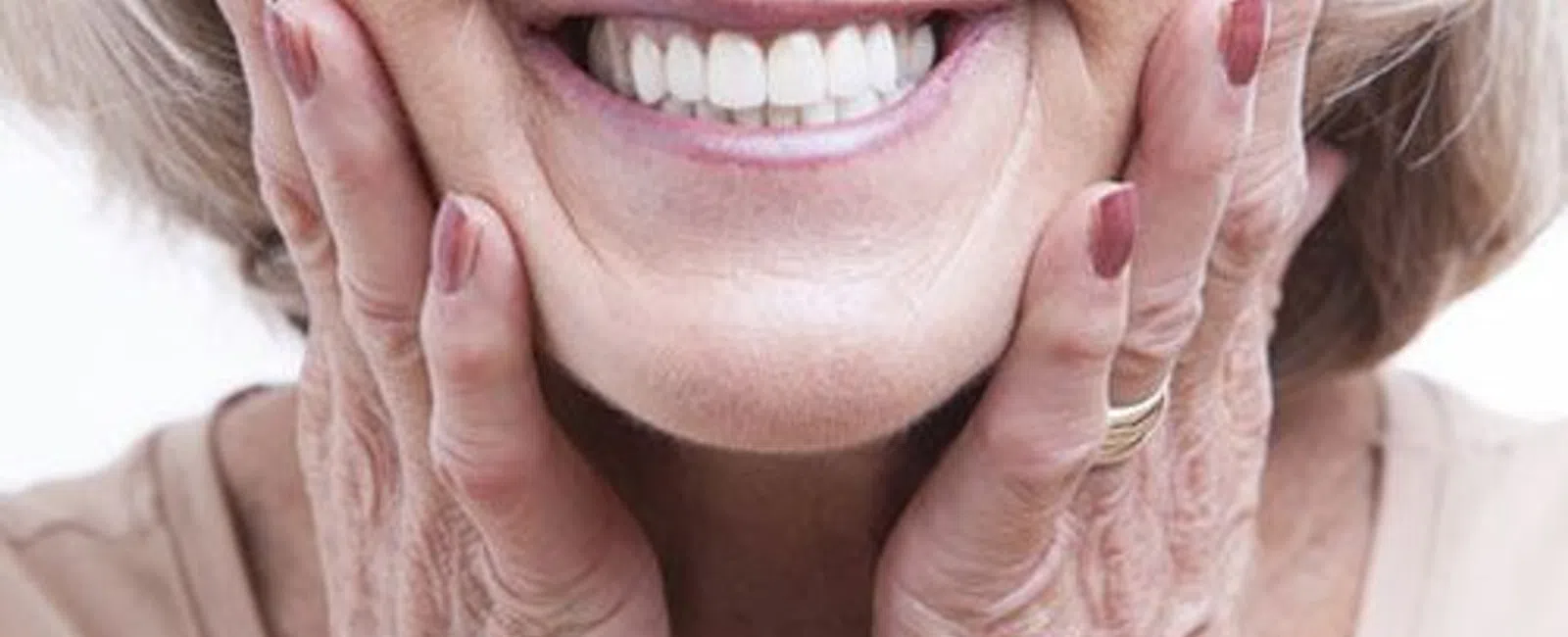 Close-up of an elderly woman smiling with hands gently on her cheeks showing white teeth and natural wrinkles
