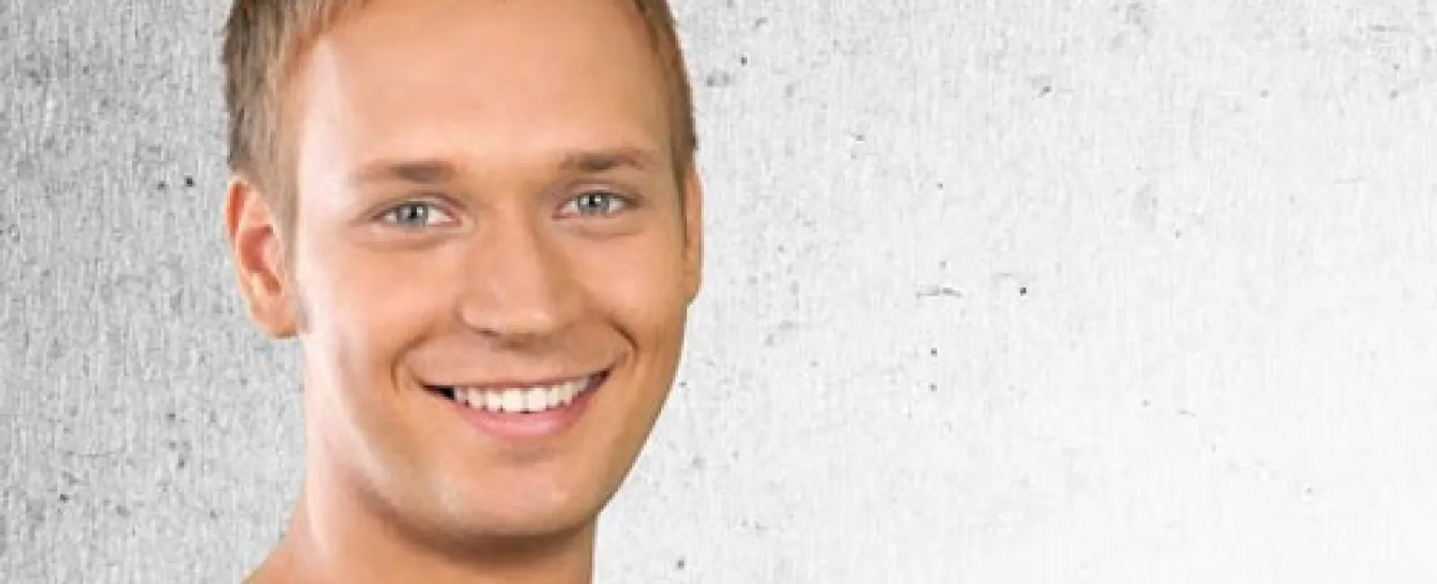 Smiling young man with short hair wearing a gray shirt standing against a textured white and gray wall.