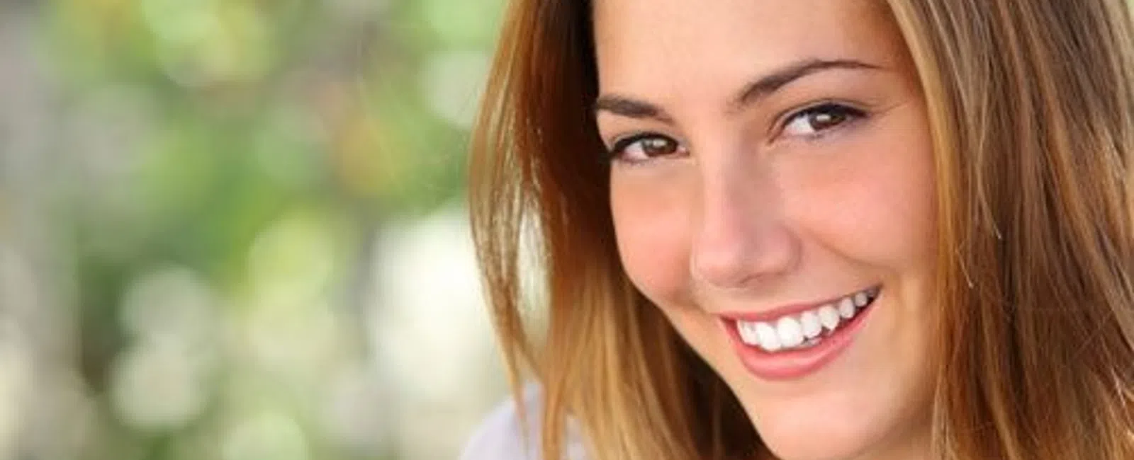 Young woman with long brown hair smiling warmly against a blurred green nature background.