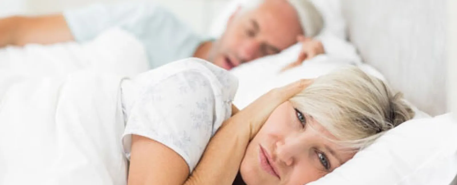 Woman lying awake covering ears while man sleeps beside her in bed, indicating noise disturbance or insomnia.