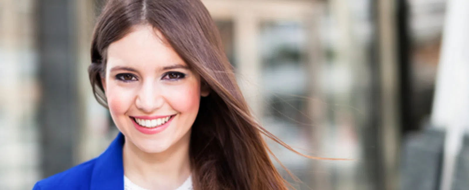 Smiling young woman with long brown hair wearing blue blazer and white lace top outdoors in daylight