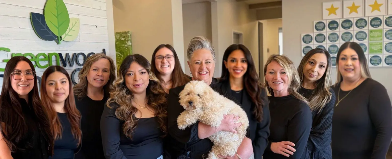 Group of smiling women wearing black tops posing indoors with a fluffy white dog in front of a Greenwood sign.