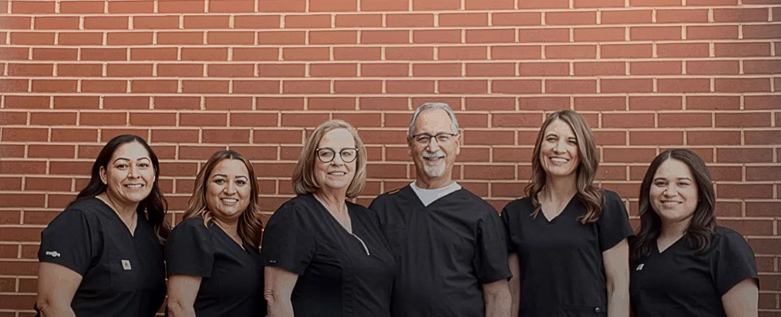 Smiling medical team in black scrubs standing against a brick wall background in a group portrait.