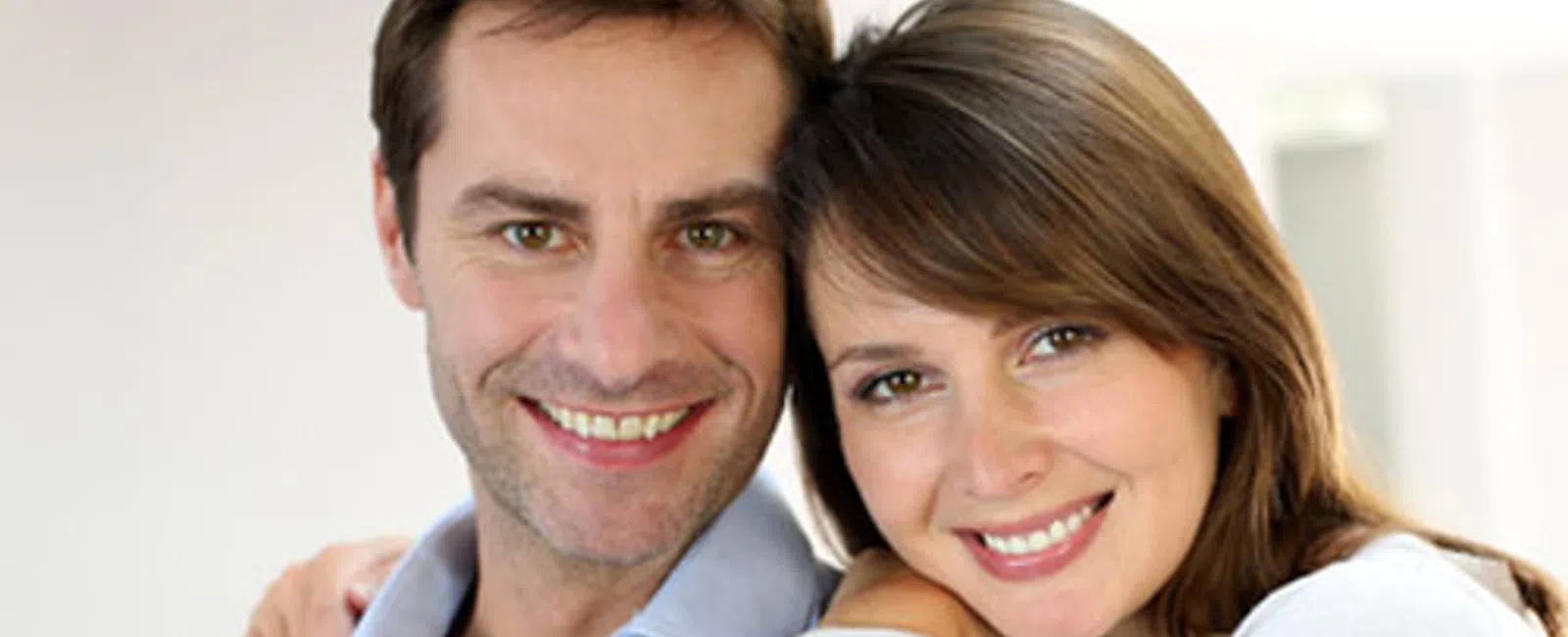 Smiling couple embracing indoors with bright background, showing affection and happiness in casual clothing.